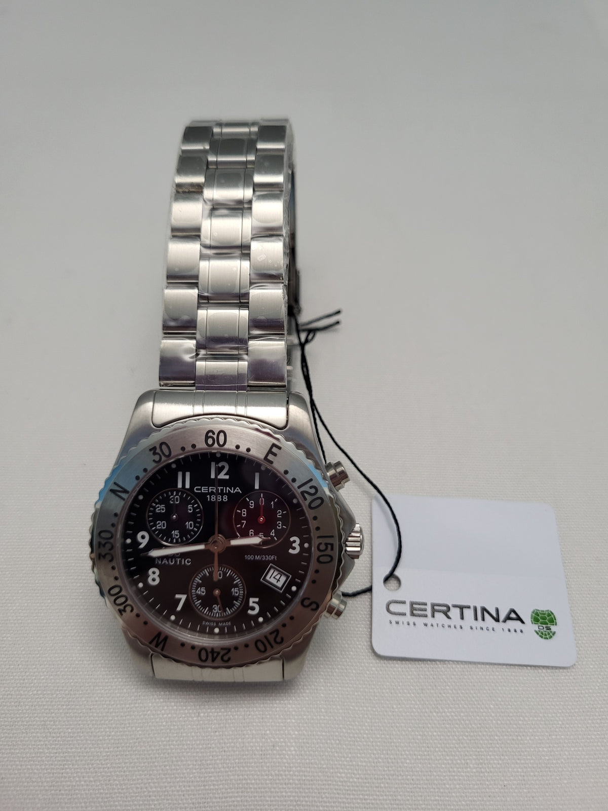 Certina automatic chronograph watch with a stainless steel link bracelet, silver-tone case, and a dark brown dial, shown with its white Certina brand tag on a light gray textured background.