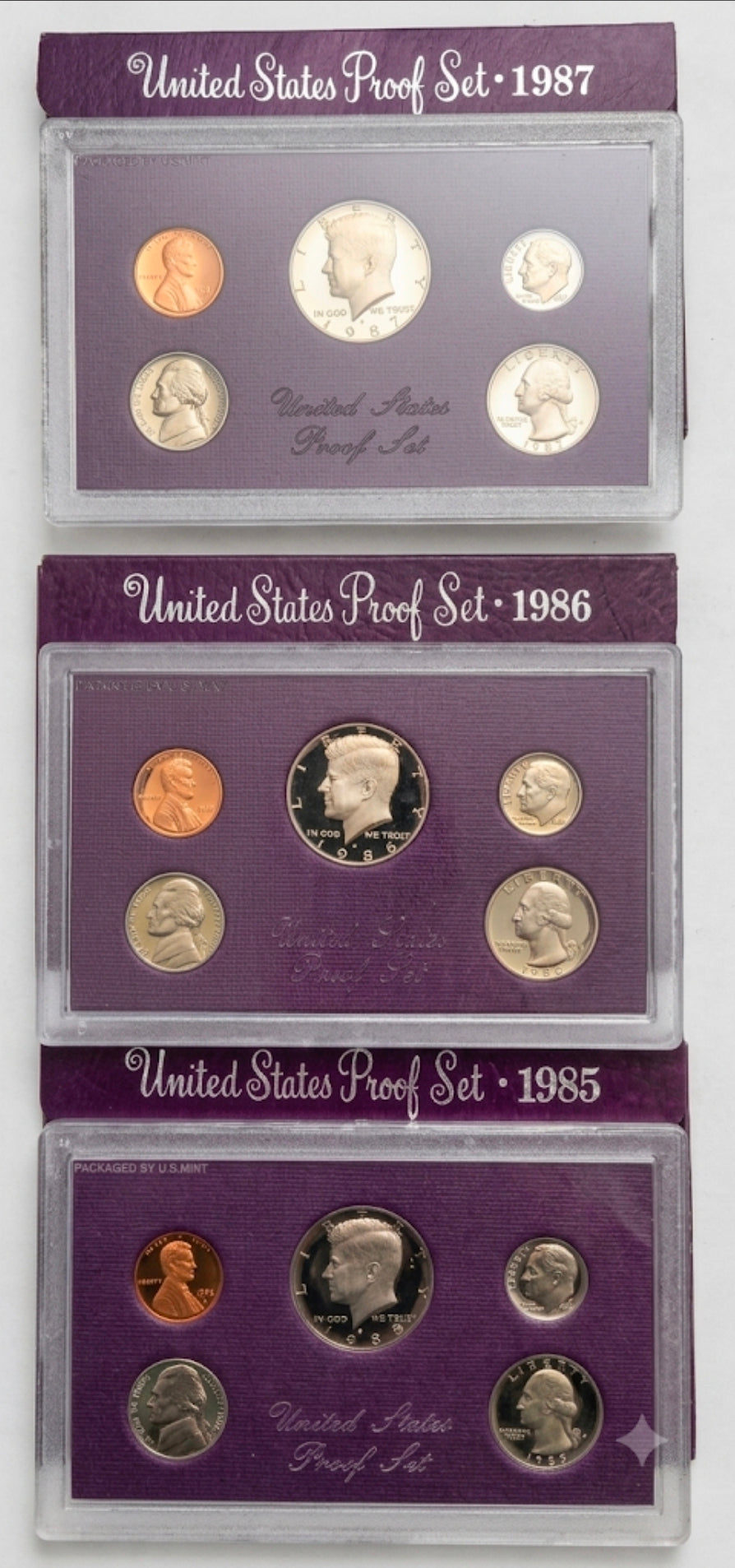 Three stacked sets of United States Proof Set coins from 1985, 1986, and 1987, each in a clear plastic holder with a purple backing card.